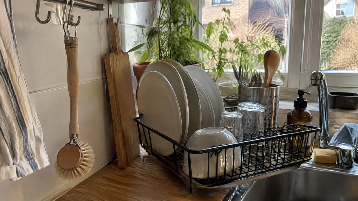 A candid phone photo of a wooden dish brush hanging beside a kitchen sink, with freshly washed plates on a drying rack a