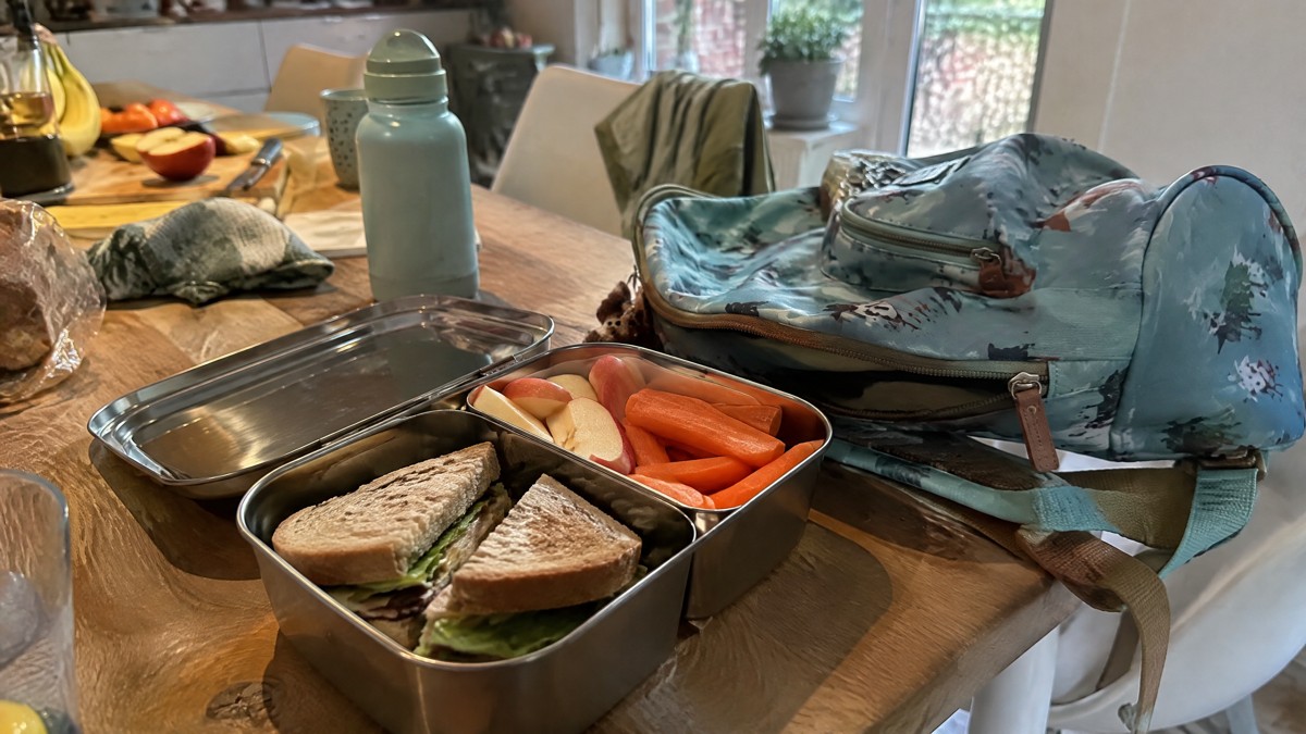 A candid phone photo of an open stainless steel lunchbox with sandwiches and sliced fruit beside a child's school bag on