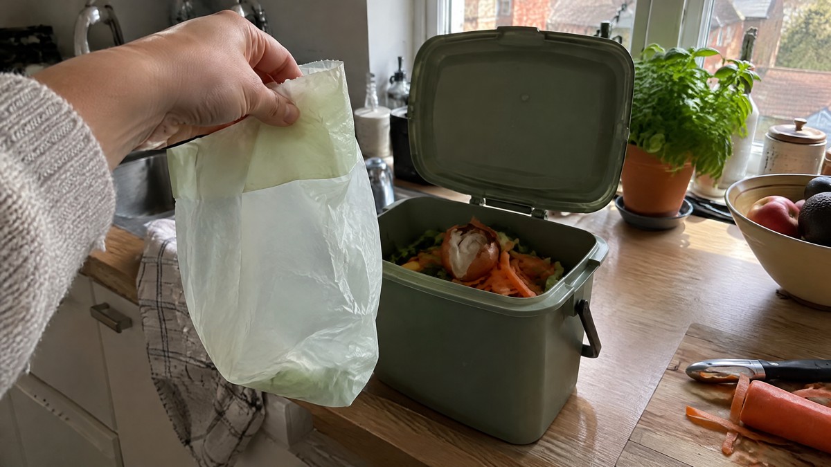 Composteerbare vuilniszak: wat is het en mag hij in je gft? A candid phone photo in a lived-in Belgian kitchen shows a hand holding an unmarked translucent compostable bin liner be