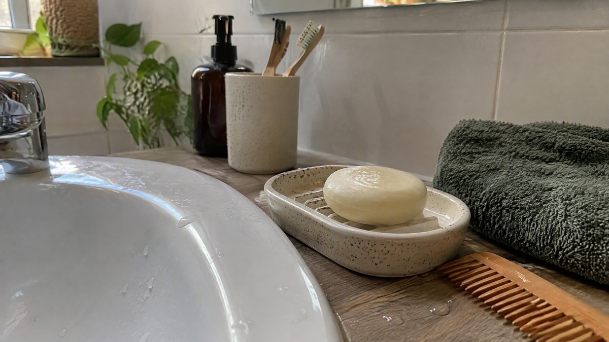 A candid phone photo of a damp conditioner bar on a draining ceramic soap dish beside a comb and a folded towel in an or
