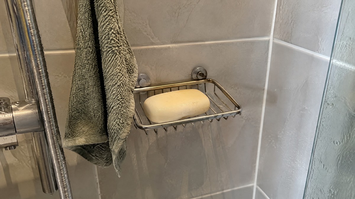 A candid phone photo of a slightly used soap bar drying on a draining soap dish in an ordinary home shower, with a damp