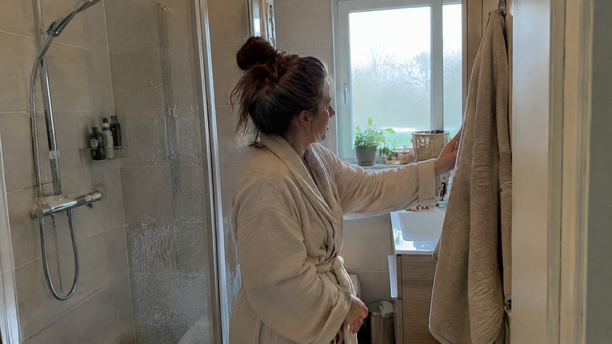 A candid phone photo in a modest home bathroom shows a person in a bathrobe reaching for a plain cotton towel beside a s