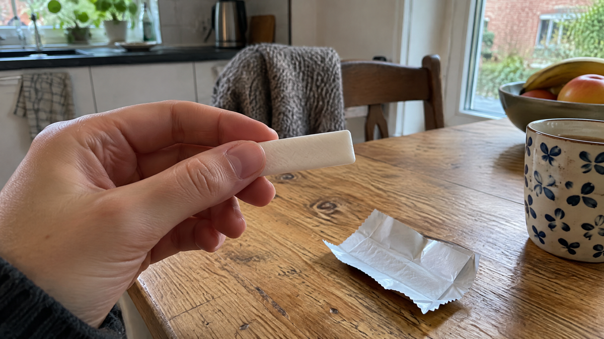 A candid phone photo of a hand holding an unwrapped piece of chewing gum above a kitchen table beside a plain folded wra