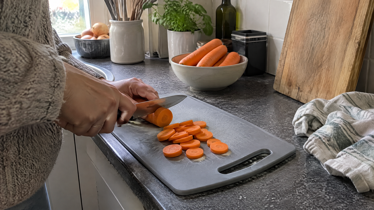 A candid phone photo in natural window light shows a hand slicing a carrot on a worn grey plastic cutting board in an ev