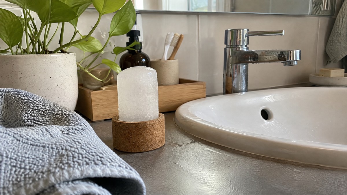 A candid phone photo in soft morning light shows a damp crystal deodorant in a cork holder beside a bathroom sink, with