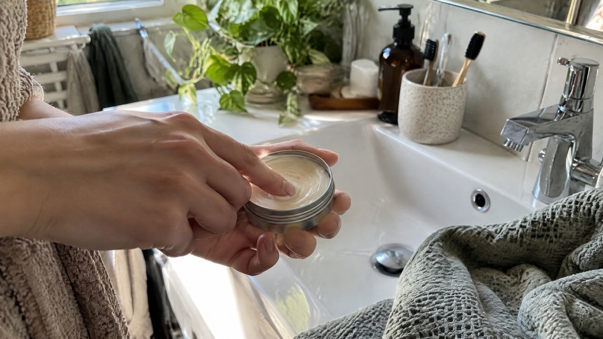 A candid phone photo in soft morning light shows a hand taking a small dab of deodorant cream from an unlabelled tin bes
