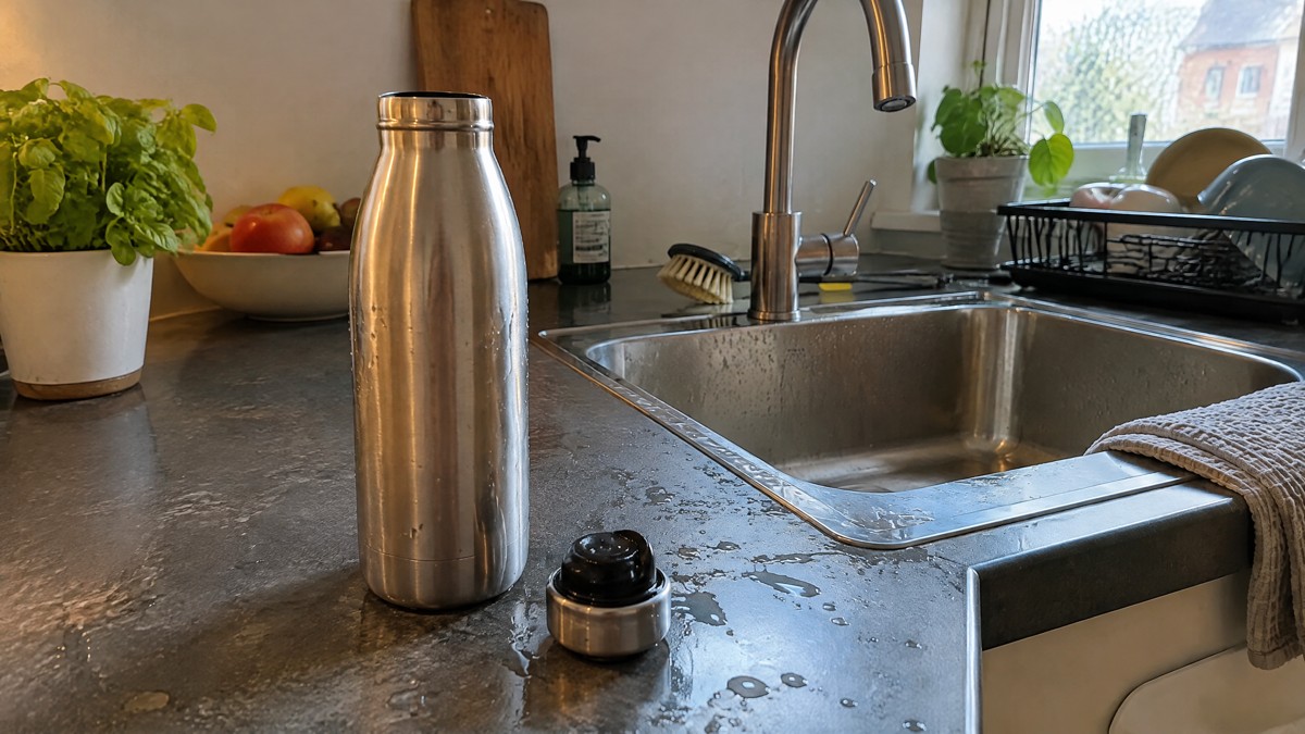 A candid phone photo of a reusable stainless steel water bottle with its cap beside it on a slightly wet kitchen counter
