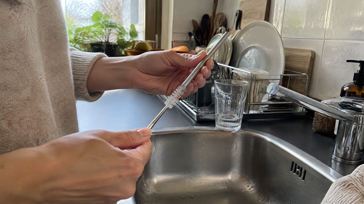 A candid phone photo of hands cleaning a reusable stainless steel straw with a small brush over an ordinary kitchen sink