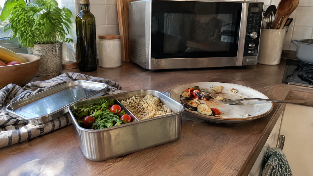 A candid phone photo in a lived-in home kitchen shows an open stainless steel lunchbox beside a ceramic plate of leftove