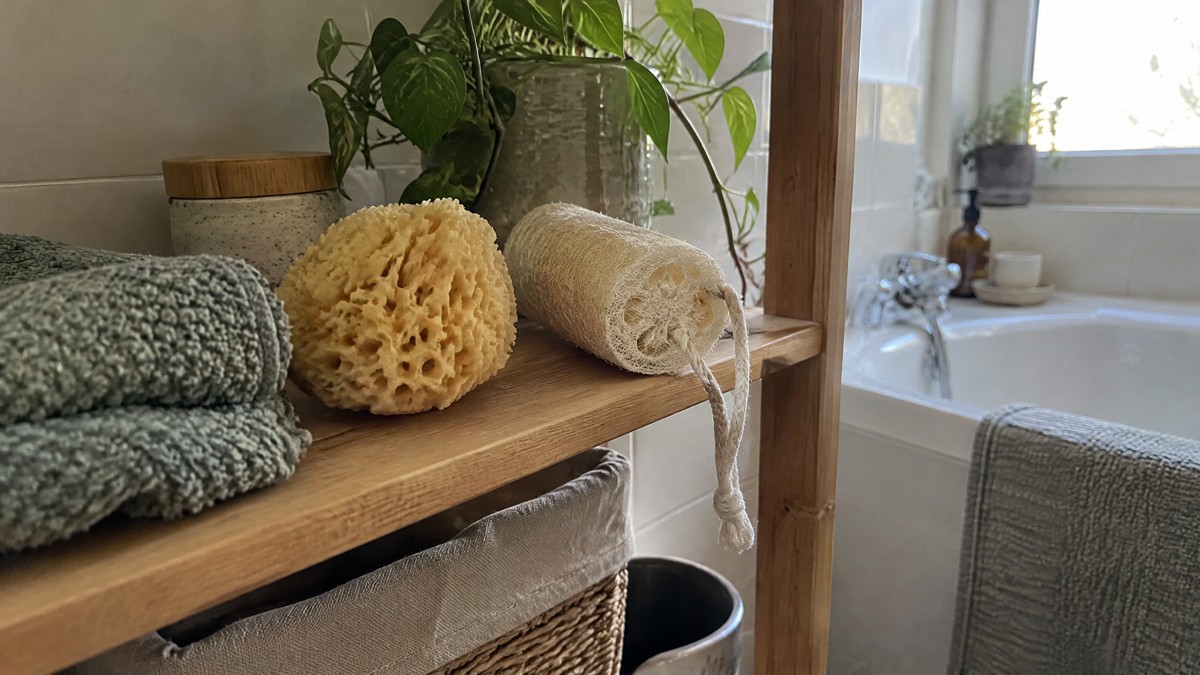A candid phone photo in a modest home bathroom shows a natural sea sponge and a dry loofah on an open wooden shelf besid