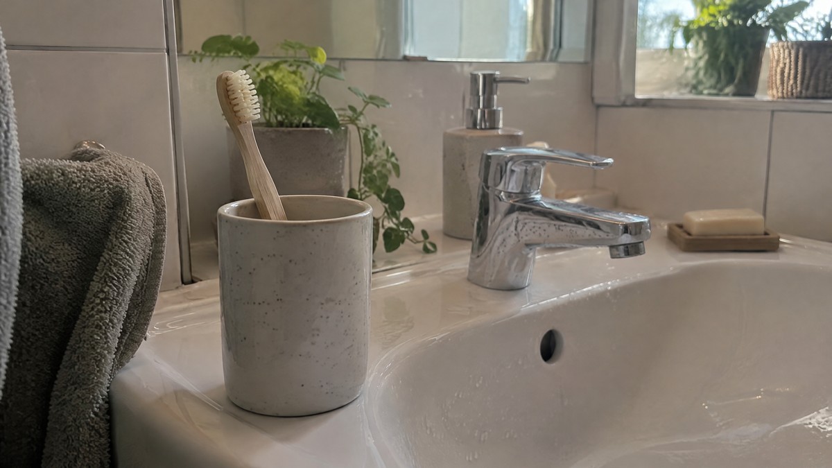 A candid phone photo of a wooden toothbrush with natural bristles standing upright in an open ceramic cup beside a sligh